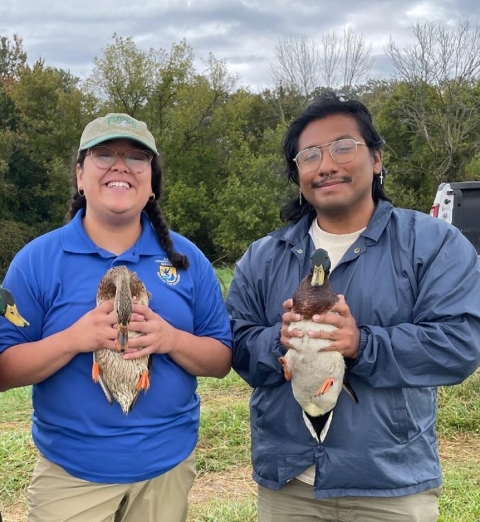 two young people wearing glasses smile while holding mallards in their hands. Forest is seen behind them